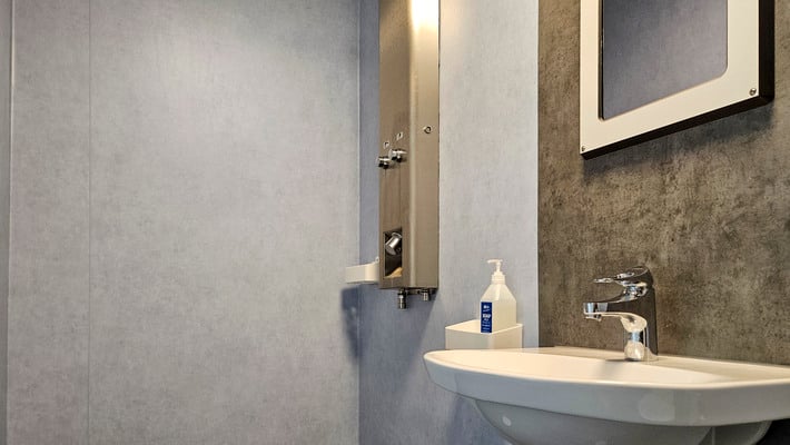 Toalett med gråa väggar. Handfat med spegel och duschmunstycke.