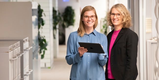 Två medarbetare på Locum står bredvid varandra i en korridor och håller i en Ipad och tittar mot oss. 